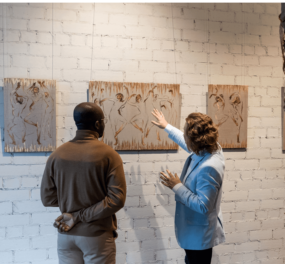 Two people discussing paintings on brick wall.